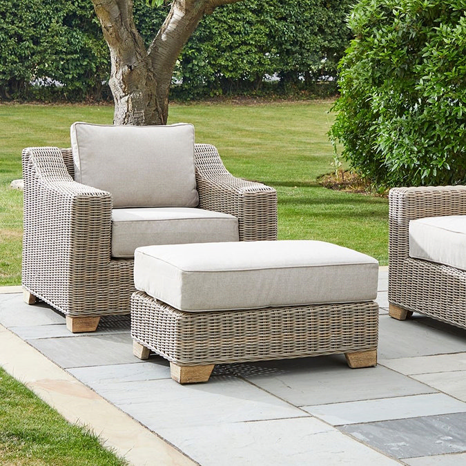 image of beige rattan footstool with plush cushion, in front of matching armchair on patio