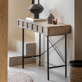 image of smoked oak console table with panelled drawers and black metal underframe, positioned against a hallway wall next to some stairs with a small woven rug in front of it, and topped with a candle.