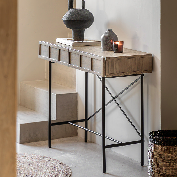 image of smoked oak console table with panelled drawers and black metal underframe, positioned against a hallway wall next to some stairs with a small woven rug in front of it, and topped with a candle.