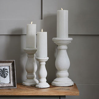 image of trio of matt white pillar candle holders with curved column-inspired  bases, stood on a side table holding battery-operated candles