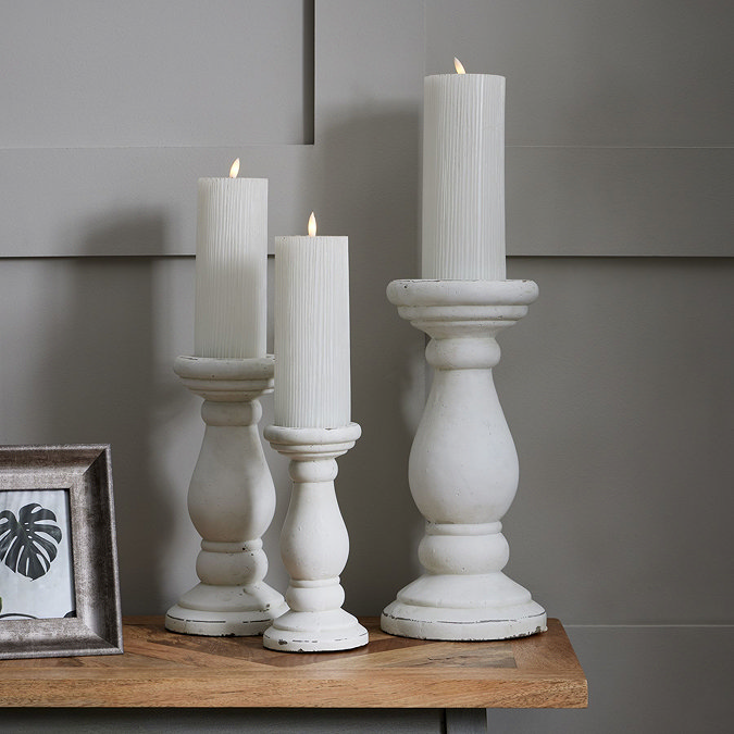 image of trio of matt white pillar candle holders with curved column-inspired  bases, stood on a side table holding battery-operated candles