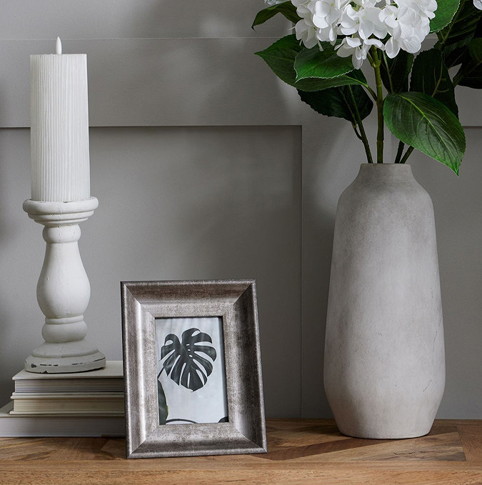 image of small matt white ceramic candle holder in column-inspired design, on top of three books next to a picture frame and stone vase containing a white flower, on top of an oak sideboard