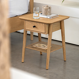 light oak bedside table with one drawer on light grey floor. book and glass of water resting on top, with book on lower shelf