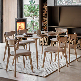 rectangular wooden table in a light oak with six chairs around it, in a dining room with rug underneath and lit fire in the corner