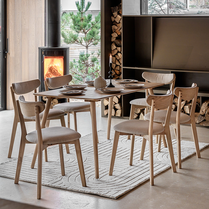 rectangular wooden table in a light oak with six chairs around it, in a dining room with rug underneath and lit fire in the corner