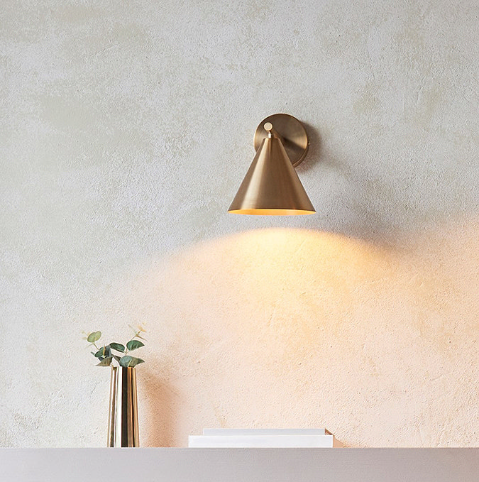 image of brass coloured wall light above a living room shelf, emitting warm glow onto thin faux plant