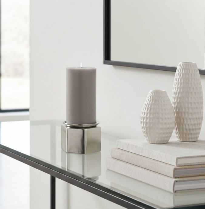 image of grey pillar candle in metal geometric candle holder on top of glass sideboard, next to pile of cream books and decorative accessories