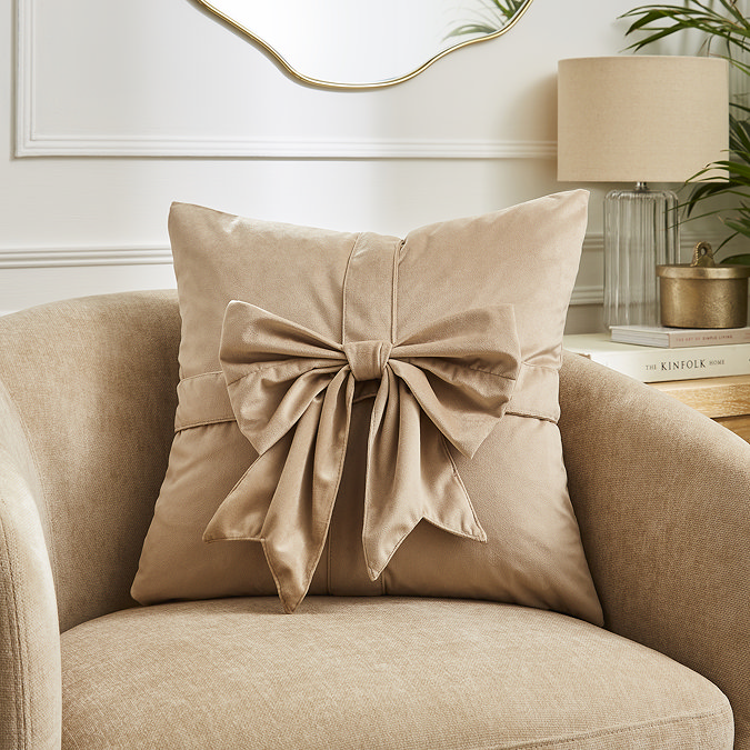 image of natural coloured square cushion with velvet bow on front, sat on a cream armchair