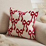 image of 30x30cm natural coloured cushion with velvet red bow pattern, sat in the corner of a cream armchair