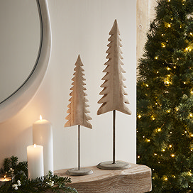 medium whitewashed nordic tree on stand, next to smaller version of the same product on a light oak mantlepiece with christmas tree in background