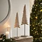 medium whitewashed nordic tree on stand, next to smaller version of the same product on a light oak mantlepiece with christmas tree in background