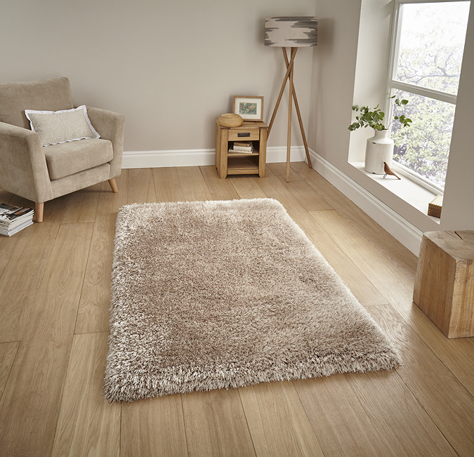 image of large beige shaggy rug in the middle of wooden flooring in front of window with beige armchair to the left