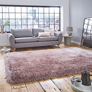 image of large pink shaggy rug in front of light grey sofa with pink accent cushions on rustic wooden floor, backlit by big apartment windows