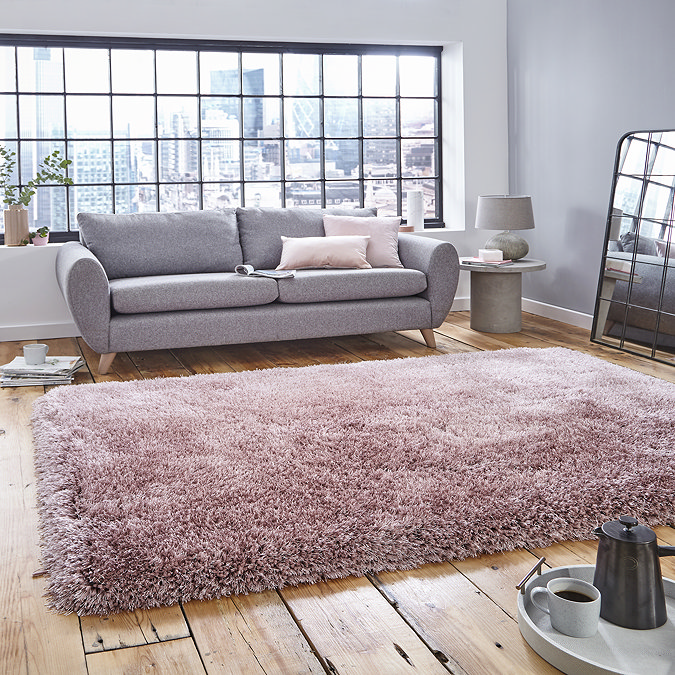 image of large pink shaggy rug in front of light grey sofa with pink accent cushions on rustic wooden floor, backlit by big apartment windows