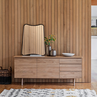 image of oak sideboard with two wide doors and three horizontal drawers, with large wavy mirror and faux plant on top, pushed against a wooden slatted wall