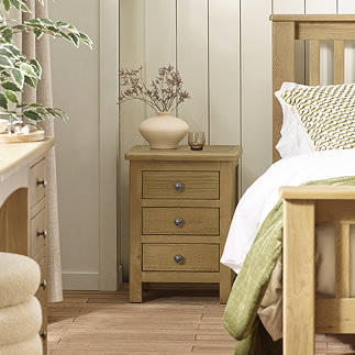 image of solid oak three drawer bedside, with round metal handles and decor on top of table, next to matching bed