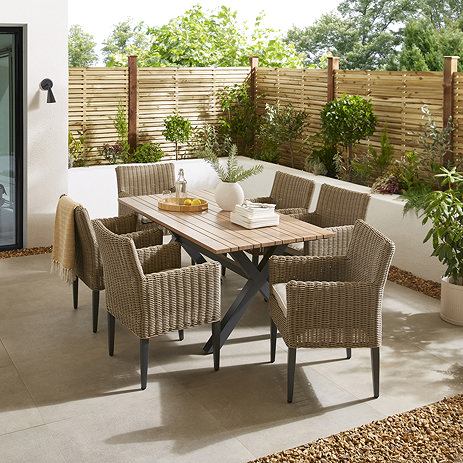 image of wooden rectangular table with six rattan chairs tucked around it, with black metal legs, on stone patio of garden surrounded by fence and foliage