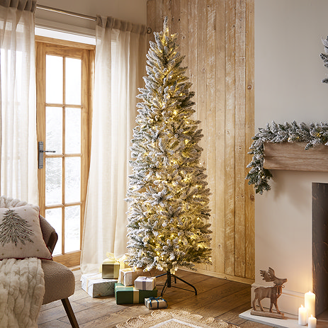 pre-lit slim snowy spruce tree in middle of warmly lit living room