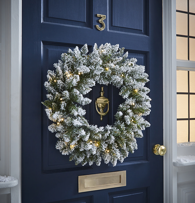 snowy spruce wreath, lit with warm white LEDs, hung on a navy blue front door at night