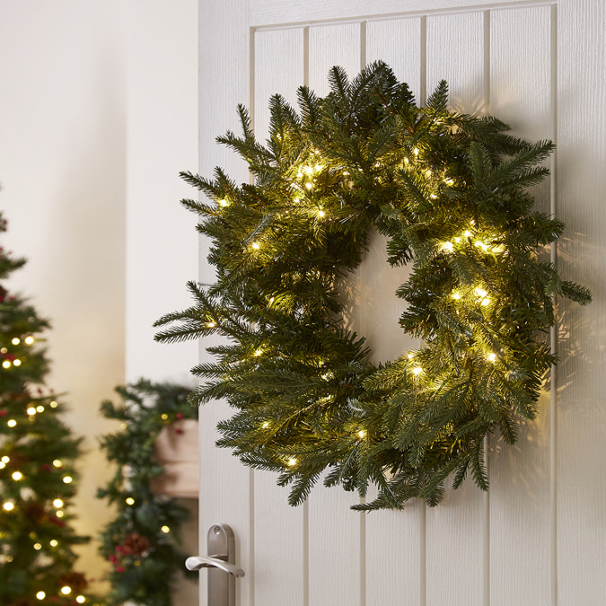 pre-lit dark green wreath with 100 LEDs on the back of a white living room door, with christmas tree and garland in the background