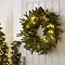 pre-lit dark green wreath with 100 LEDs on the back of a white living room door, with christmas tree and garland in the background