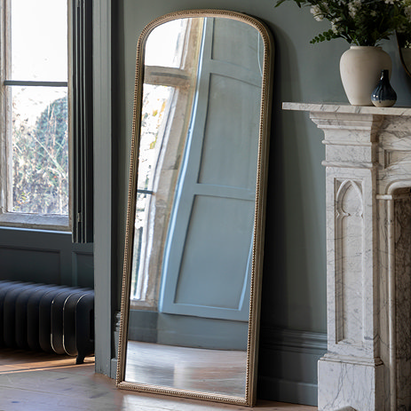 image of large rectangular mirror rounded at top, leaning against pale green wall next to marble fireplace