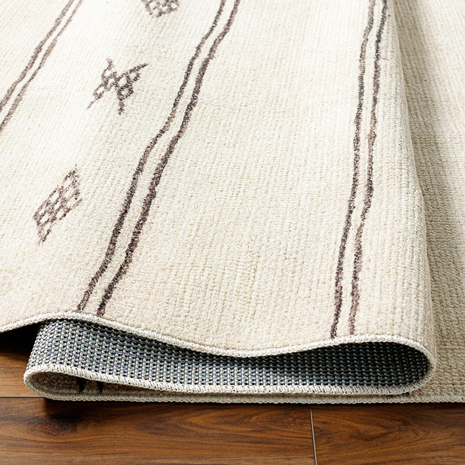 close up image of ivory rug with brown scandi pattern design, ruched up and folded slightly on wooden flooring to show its thinness