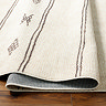 close up image of ivory rug with brown scandi pattern design, ruched up and folded slightly on wooden flooring to show its thinness