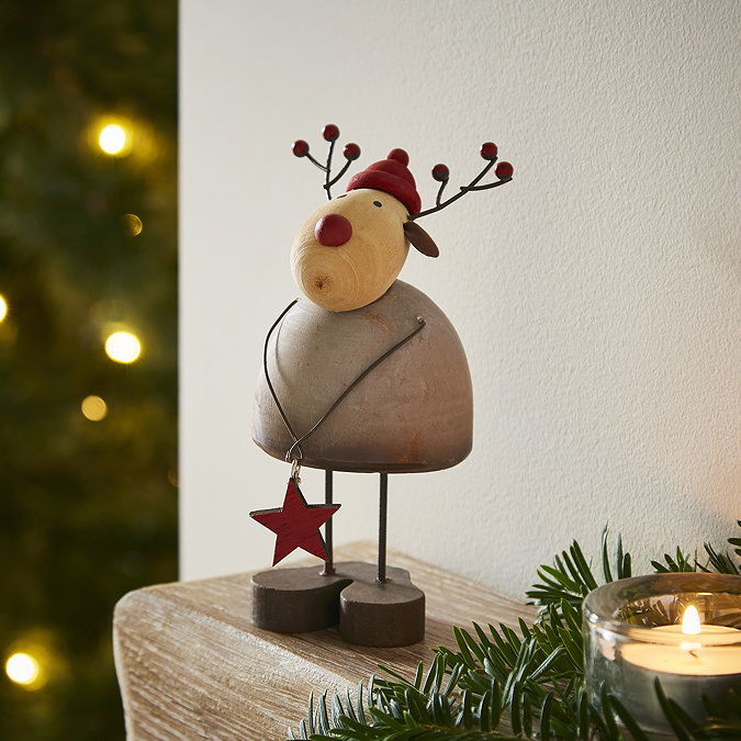 rustic wooden reindeer with red hat and nose detailing, holding small red wooden star, standing on the edge of an oak wood mantlepiece with garland and candle.
