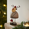rustic wooden reindeer with red hat and nose detailing, holding small red wooden star, standing on the edge of an oak wood mantlepiece with garland and candle.