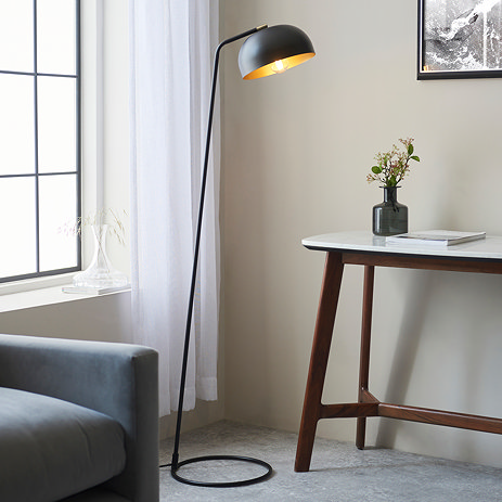 image of matt black thin floor lamp with curved head and domed shade next to grey sofa and modern brown console table