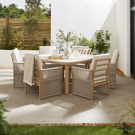 image taken at mid-level of luxury rope seats with cream cushions, around a cream sintered stone triangular curved table, on garden patio surrounded by fences and foliage