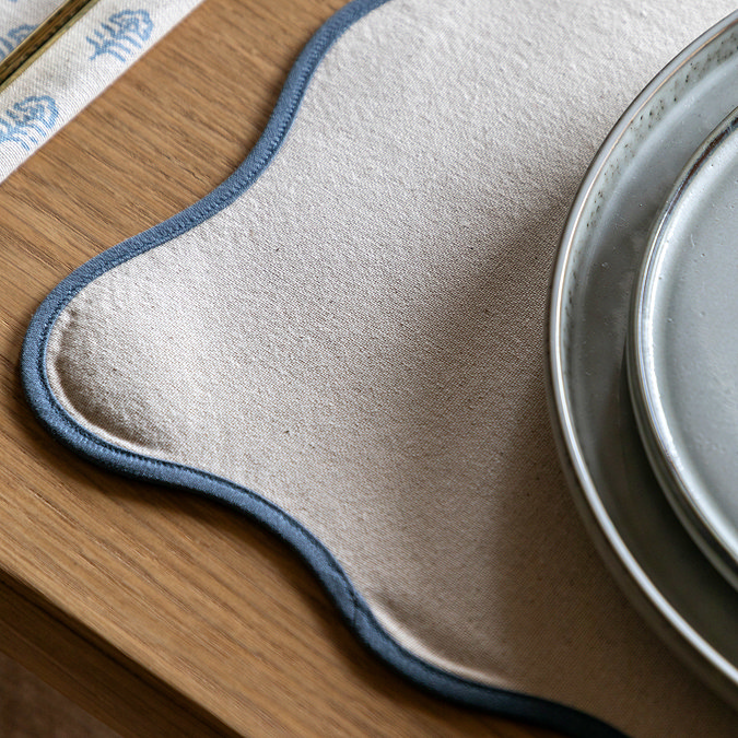 close up image of cream placemat with blue scalloped edge, dinnerware sitting on top of it