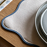 close up image of cream placemat with blue scalloped edge, dinnerware sitting on top of it