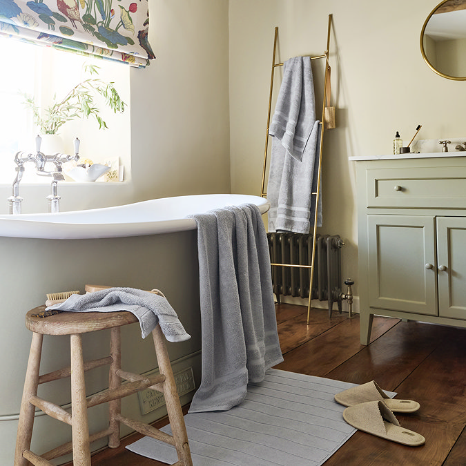 wide shot of bathroom with light grey towels draped over the bath, on a stool, hung up on a wall, with coordinating bath mat on the floor