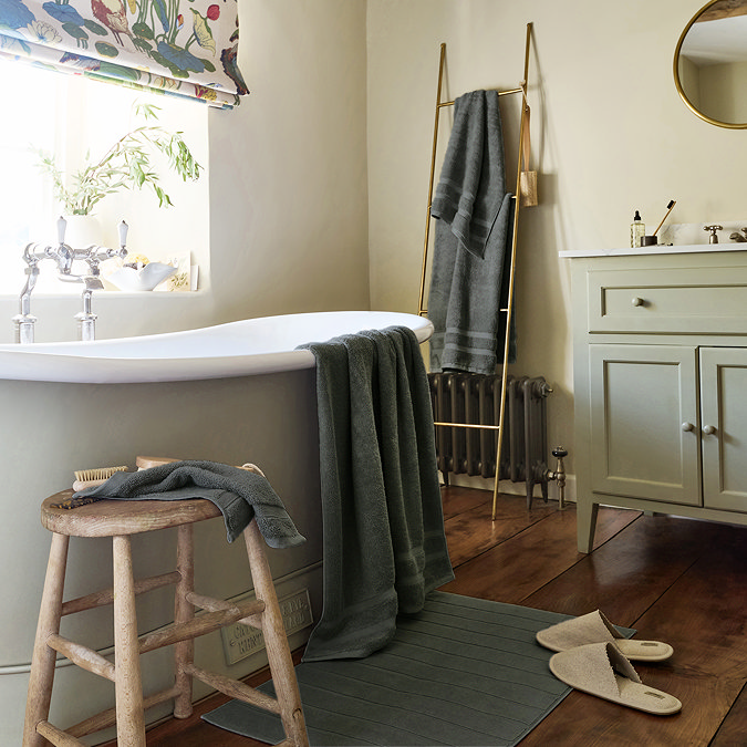 wide image of bathroom with bath sheet on side of the bath, hand towel on a small side stool, and bath towel hung up on wall with coordinating bath mat on the floor