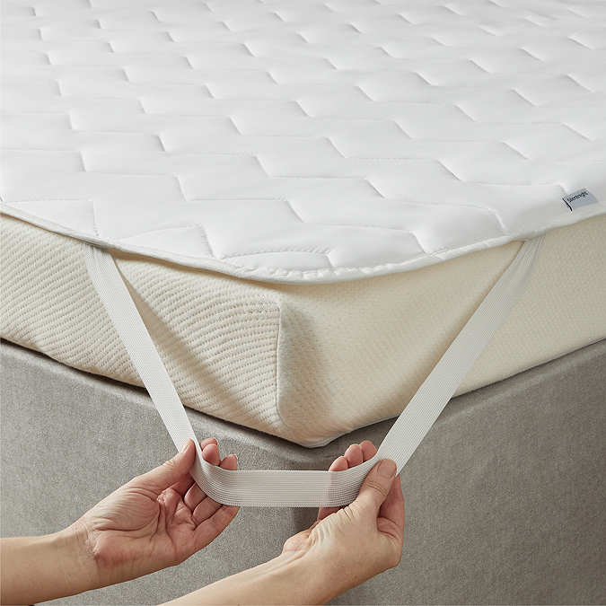 close up of a person's two hands stretching the elastic band of a mattress topper over a corner of the bed to secure it