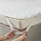 close up of a person's two hands stretching the elastic band of a mattress topper over a corner of the bed to secure it