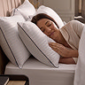 side on image of woman resting in bed with head on pair of hotel pillows, white with blue edging