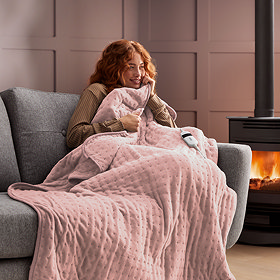 image of ginger woman on grey sofa next to a lit fire, wrapped in soft pink heated throw