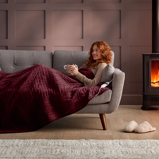 image of ginger woman reclining sideways on grey sofa, wrapped up in burgundy red heated throw
