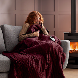 ginger woman on a sofa next to a lit fire, wrapped in a burgundy red heated blanket