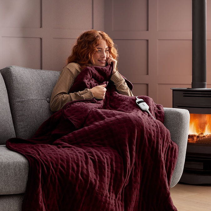 ginger woman on a sofa next to a lit fire, wrapped in a burgundy red heated blanket