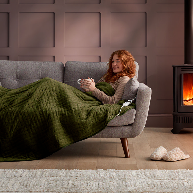 image of woman reclining on sofa holding a mug, covered in olive green heated throw