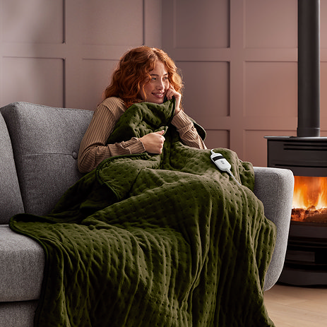 image of ginger woman on a grey sofa next to a lit fire, wrapped up in olive green heated throw
