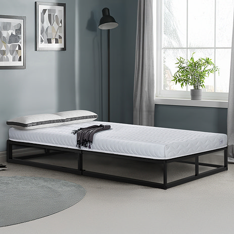 image of basic double mattress on black metal bedframe pushed against light grey wall with light streaming through bedroom wall