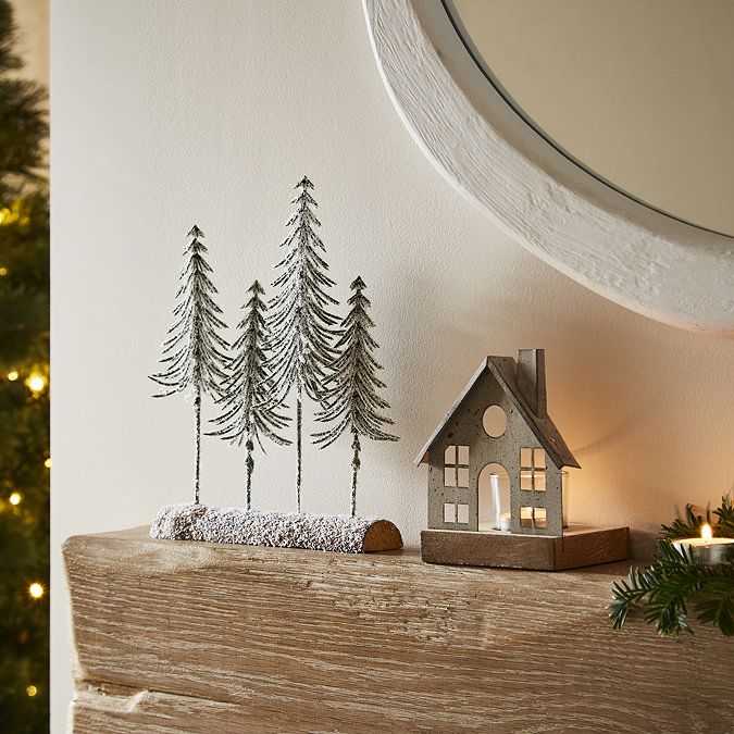 four snow-dusted trees in a wooden log, stood next to a metal tealight holder with glass votive, on an oak mantlepiece.