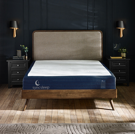 image of double mattress on wooden bed in front of navy blue bedroom wall, with syncsleep logo embroidered on the front left side