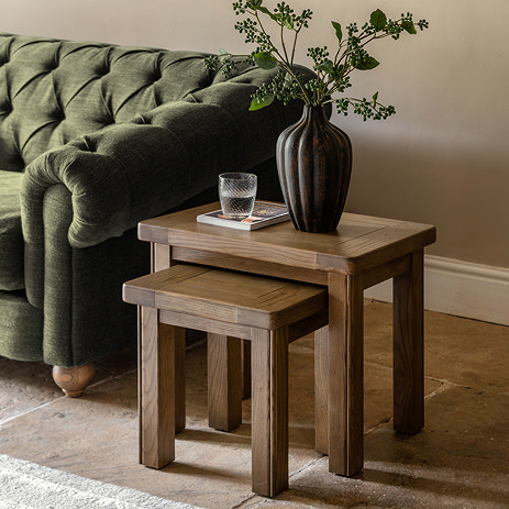 image of two dark oak nesting tables with a faux plant and glass of water on top, next to a forest green velvet sofa