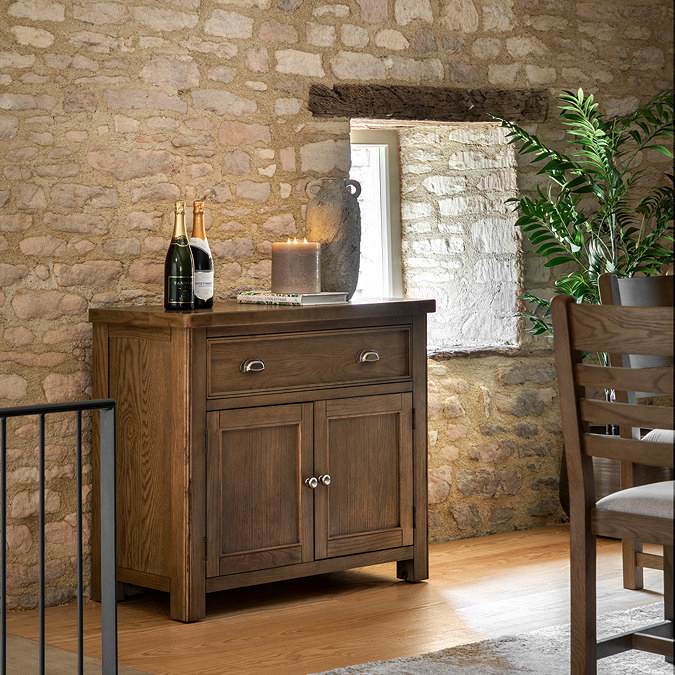 image of dark oak sideboard with one drawer and 2 doors, topped with glasses and pushed against an exposed brick wall